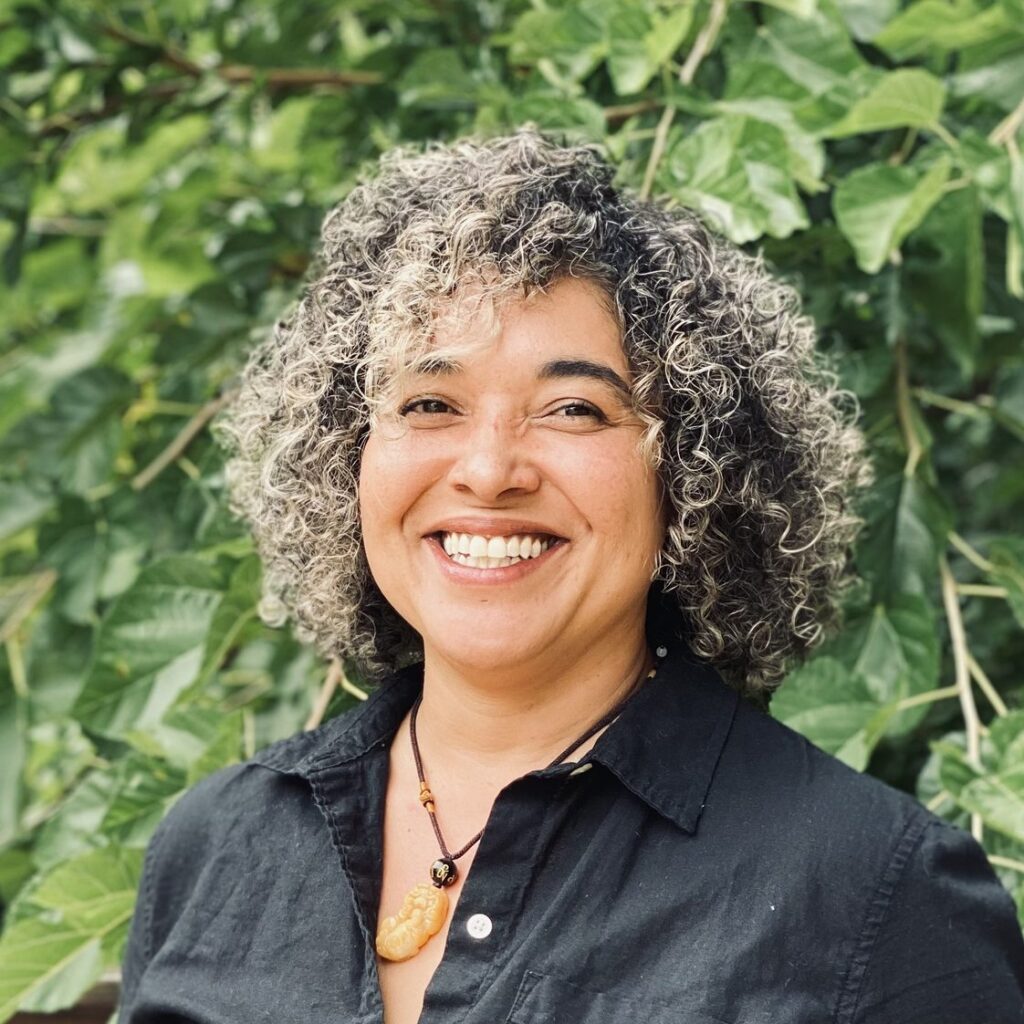 Headshot of a woman with curly hair in front of a leafy green tree